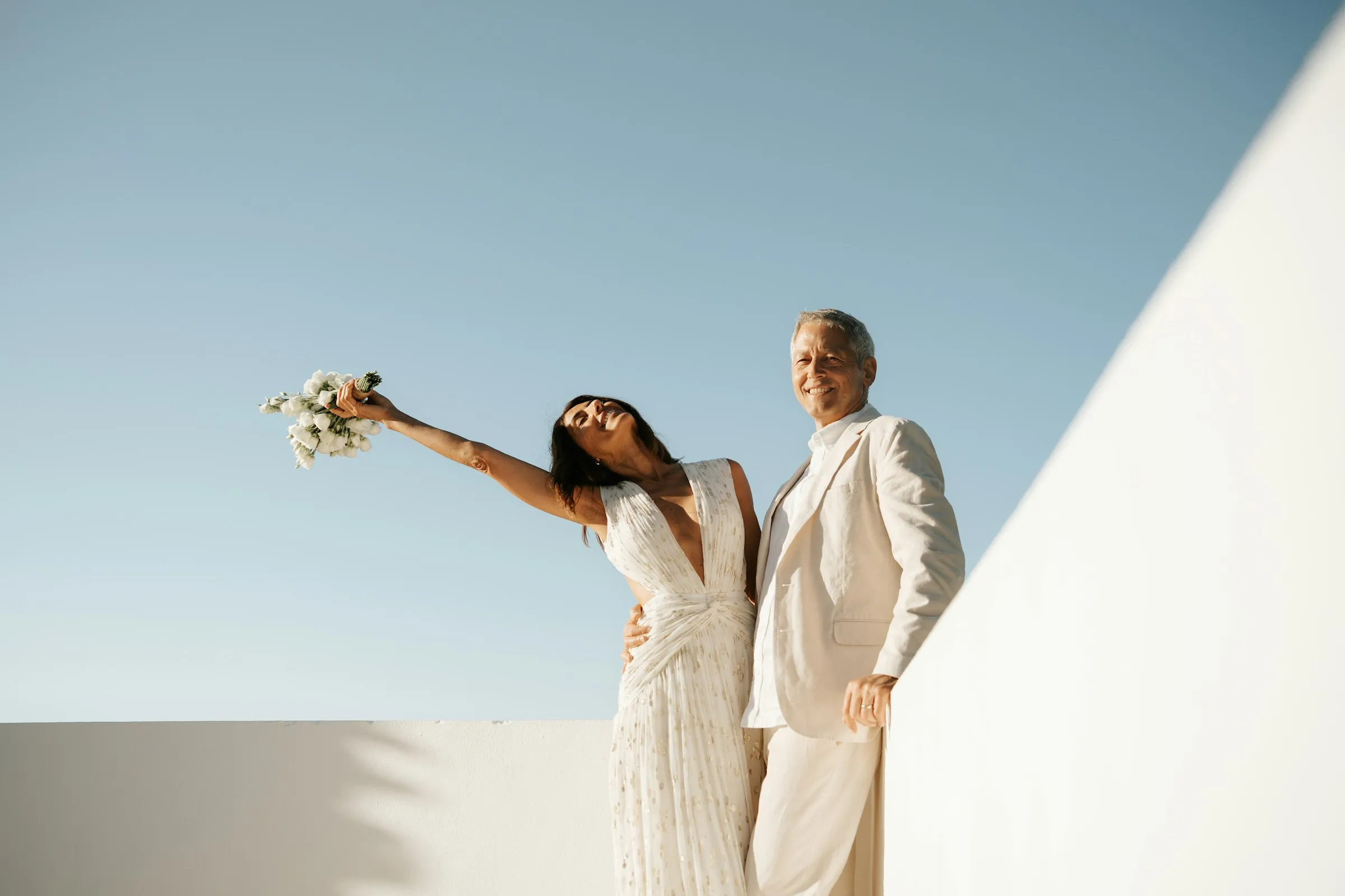 Maria raising her bouquet against a white wall and blue sky, João beside her