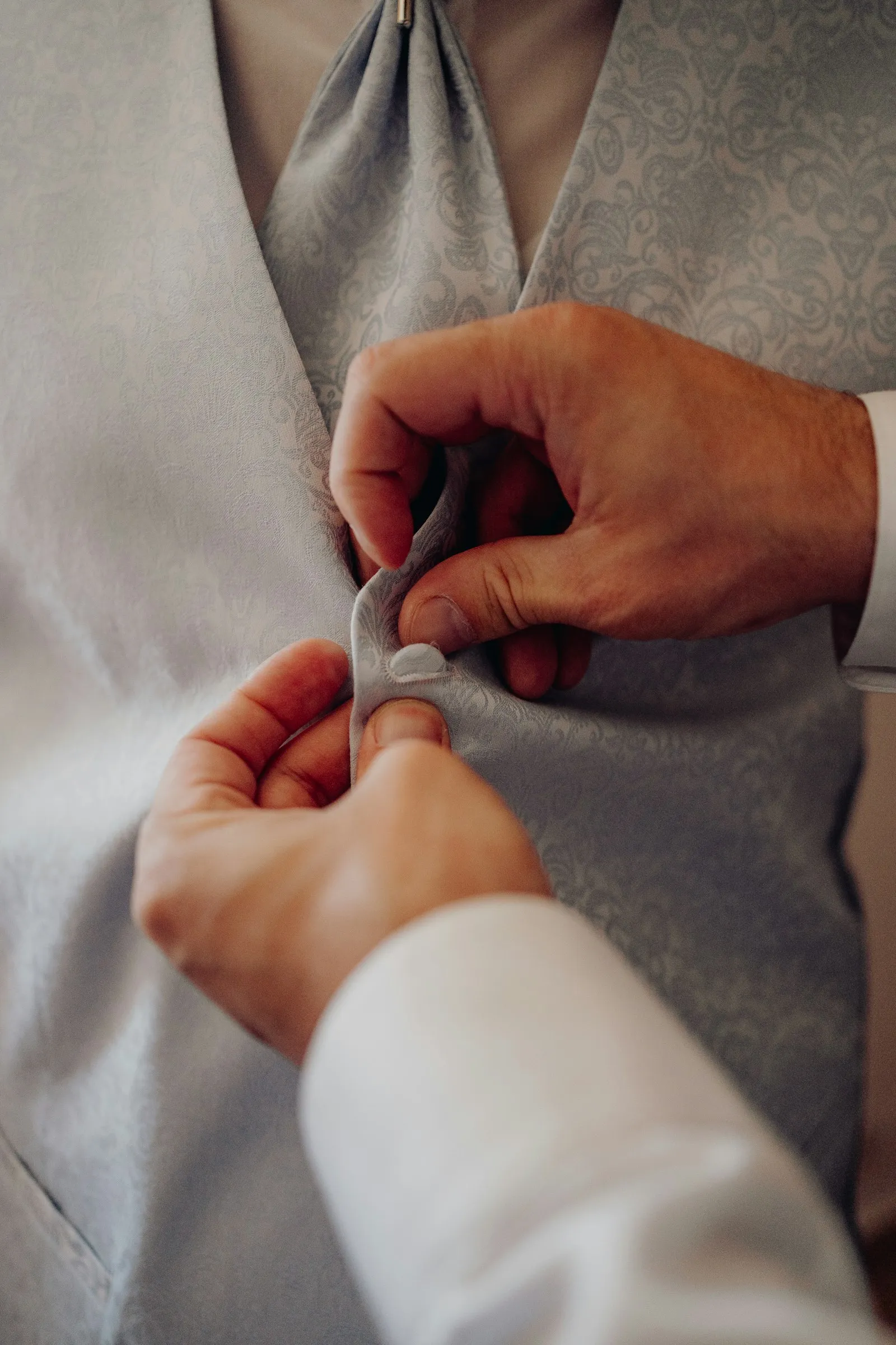 Rafael’s hands buttoning a pale grey waistcoat